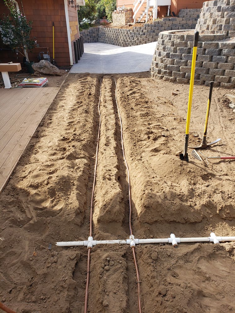 Trenches with subsurface drip irrigation lines running through bare soil beside a deck, before a new sod lawn was installed