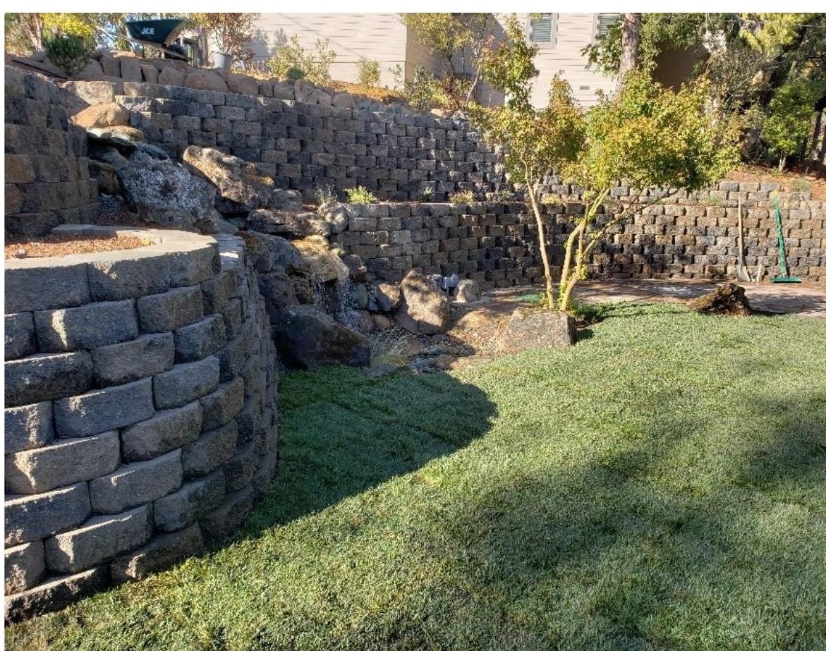 A transformed hillside front yard with stacked stone retaining walls, a natural-looking waterfall spilling into boulders, new lawn, and an ornamental tree